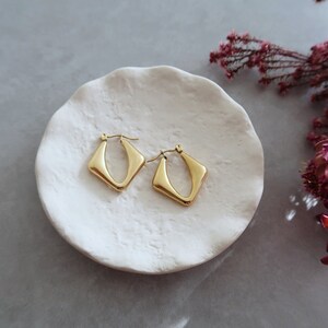 Può includere: Un paio di orecchini a cerchio dorati con un design geometrico a forma di diamante. Gli orecchini sono esposti su un piatto in ceramica bianca.