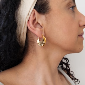 Puede incluir: Pendientes de aro dorados con un diseño texturizado y orgánico. Los pendientes se llevan en la oreja de una persona con cabello oscuro y una diadema color crema. Los pendientes tienen un patrón único y detallado.
