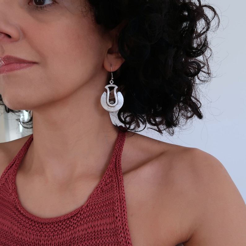 May include: Silver earrings with a unique geometric design. The earrings are shaped like a circle with a cutout in the center. The earrings are hanging from a person's ear.