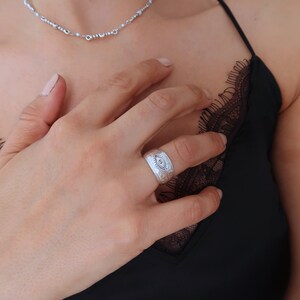 Peut inclure: Une large bague argent&eacute;e avec une texture martel&eacute;e et un motif d'&oelig;il grav&eacute; au centre entour&eacute; de lignes rayonnantes. L'anneau pr&eacute;sente une finition mate et est port&eacute; au doigt, mettant en &eacute;vidence le dessin d&eacute;taill&eacute; du symbole de l'&oelig;il.