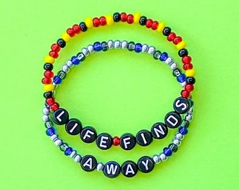 May include: Two beaded bracelets with the words "LIFE FINDS A WAY" spelled out in black beads. The bracelets are layered, with a red, yellow, and blue beaded bracelet on top of a silver beaded bracelet.