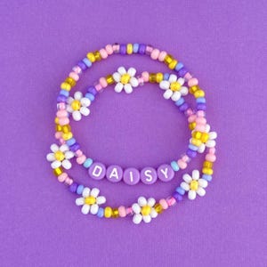 Peut inclure: Un bracelet en perles de couleur violet, jaune, rose et blanc avec le mot "DAISY" écrit en perles violettes.