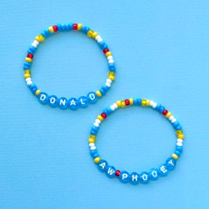 Peut inclure: Deux bracelets en perles avec les noms "DONALD" et "AW PHOOEY" écrits en lettres blanches sur des perles bleues. Les bracelets sont faits de perles bleues, blanches, jaunes et rouges.