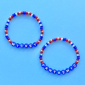 Peut inclure: Deux bracelets en perles avec des perles rouges, blanches et dorées. Le premier bracelet épelle "MICKEY" en lettres blanches sur des perles bleues. Le deuxième bracelet épelle "FANTASTIC" en lettres blanches sur des perles bleues.