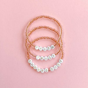 May include: Four beaded bracelets with the words "FAITH", "TRUST", "CHURRO", and "DUST" spelled out in gold lettering on white beads. The bracelets are made with light brown beads and are stacked on top of each other.