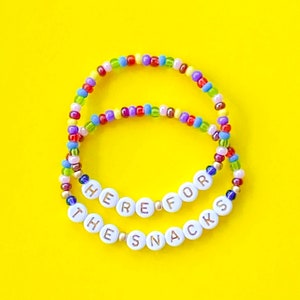 May include: Two colorful beaded bracelets with the words "Here for the Snacks" spelled out in white beads with gold lettering.