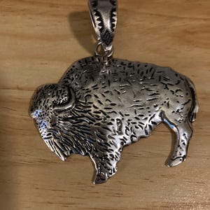 May include: A silver-toned metal pendant shaped like a buffalo in profile. The surface features textured detailing to represent fur and muscle structure. A decorative bail with etched geometric patterns is attached to the top of the buffalo figure.