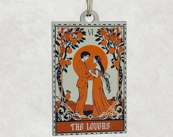 May include: A rectangular keychain featuring a tarot card design. The card depicts two figures holding hands in front of a sun, surrounded by orange trees. The text "THE LOVERS" is at the bottom, with the Roman numeral "VI" at the top.