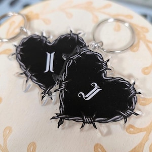 May include: Two heart-shaped keyrings. Each keyring features a black heart design with a barbed wire border. One heart has the letter "I" in white, and the other has the letter "J" in white. Silver key rings are attached.