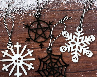 May include: A collection of gothic-themed Christmas ornaments. The ornaments include a white snowflake, a black pentagram, a black spiderweb, and a white snowflake with skull accents. Each ornament hangs from a black and white twine string.