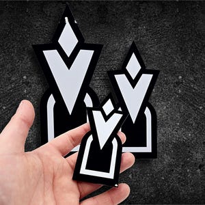 May include: Black and white geometric stickers with a stylized design. The stickers feature a diamond shape at the top, a V-shape in the middle, and a rectangular base. The stickers are in various sizes.