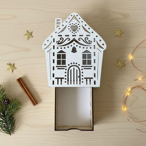 Könnte beinhalten: Eine weiße, lasergeschnittene Holzbox in Hausform mit einer offenen Schublade. Das Haus hat dekorative Ausschnitte, darunter Fenster, eine Tür und einen Schornstein. Goldene sternförmige Ornamente, ein Zimtstange und ein Tannenzweig sind um die Box angeordnet. Eine Lichterkette ist ebenfalls vorhanden.