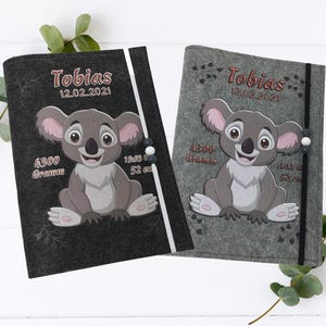Könnte beinhalten: Zwei Babybücher aus Filz, eines in Schwarz und eines in Grau, mit einem Koala-Bären-Design. Jedes Buch trägt den Namen "Tobias" und das Datum 12.02.2021 auf dem Einband, sowie das Gewicht des Babys in Gramm und die Größe in Zentimetern.