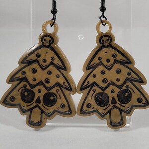 Op de afbeelding: Een paar gouden oorbellen in de vorm van kerstboom met zwarte schedelgezichten en zwarte stippen.