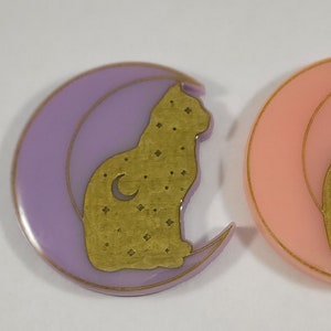 Könnte beinhalten: Zwei Harz-Anhänger mit Katze und Mondsichel. Einer ist lila und der andere rosa. Beide haben eine goldene Katzensilhouette mit einer Mondsichel und Sternen.