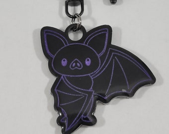 Pode incluir: Chaveiro de metal preto com um amuleto de morcego com contorno roxo. O morcego tem um rosto de desenho animado bonito com olhos grandes.