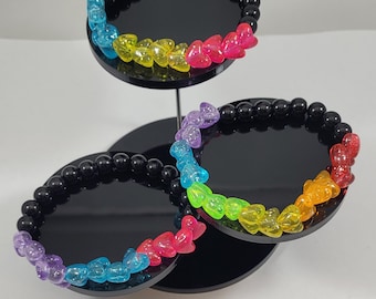 May include: Three bracelets with black beads and rainbow heart-shaped charms. The bracelets are displayed on a black tiered stand.