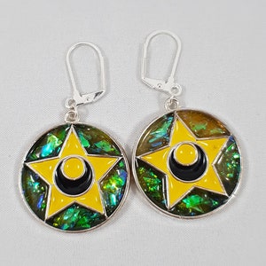 Könnte beinhalten: Ein Paar silberne Ohrringe mit einem gelben Stern- und Halbmond-Design auf einem grün schillernden Hintergrund.