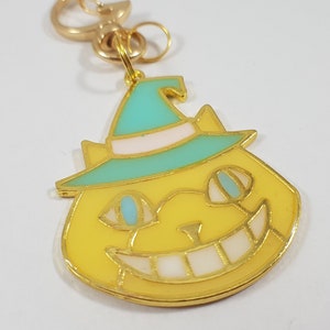 Puede incluir: Un llavero dorado con un gato de dibujos animados que lleva un sombrero de bruja turquesa con un borde blanco. El gato tiene pelo amarillo, ojos azules y una amplia sonrisa con dientes.