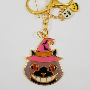 May include: A gold-tone keychain with a gray cat wearing a pink witch hat with black bats. The cat has red eyes and a white smile. The keychain also includes a small white ghost and a yellow jack-o-lantern charm.