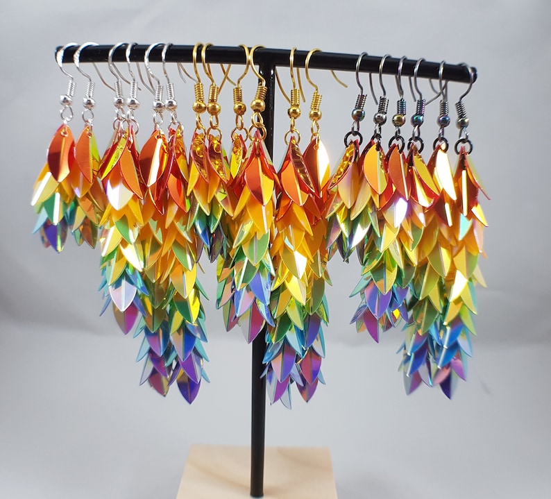 May include: A set of 10 rainbow iridescent dangle earrings with a scaly, dragon-like design. The earrings are displayed on a stand.