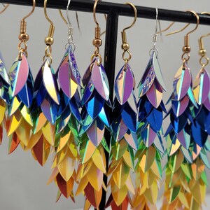 K&ouml;nnte beinhalten: Ein Paar regenbogenfarbene, schimmernde Ohrringe mit einem abgestuften Design. Die Ohrringe bestehen aus mehreren Schichten irisierender Schuppen, die einen schimmernden Effekt erzeugen. Die Ohrringe h&auml;ngen an goldfarbenen Haken.