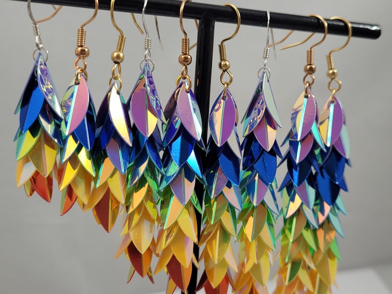 K&ouml;nnte beinhalten: Ein Paar regenbogenfarbene, schimmernde Ohrringe mit einem abgestuften Design. Die Ohrringe bestehen aus mehreren Schichten irisierender Schuppen, die einen schimmernden Effekt erzeugen. Die Ohrringe h&auml;ngen an goldfarbenen Haken.