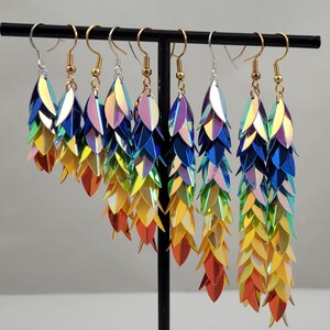 K&ouml;nnte beinhalten: Sieben Paare regenbogenfarbene, irisierende Ohrh&auml;nger mit einem abgestuften Schuppenmuster. Die Ohrringe werden auf einem schwarzen St&auml;nder pr&auml;sentiert.