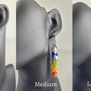 K&ouml;nnte beinhalten: Drei Paar Regenbogen-Ohrringe in verschiedenen Gr&ouml;&szlig;en: klein, mittel und gro&szlig;. Die Ohrringe sind aus Papier gefertigt und haben ein kaskadierendes Design aus bunten, schillernden Schuppen.