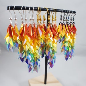 May include: Rainbow iridescent earrings with a scaly design, hanging from a black stand. The earrings are made of small, overlapping scales that create a shimmering effect.
