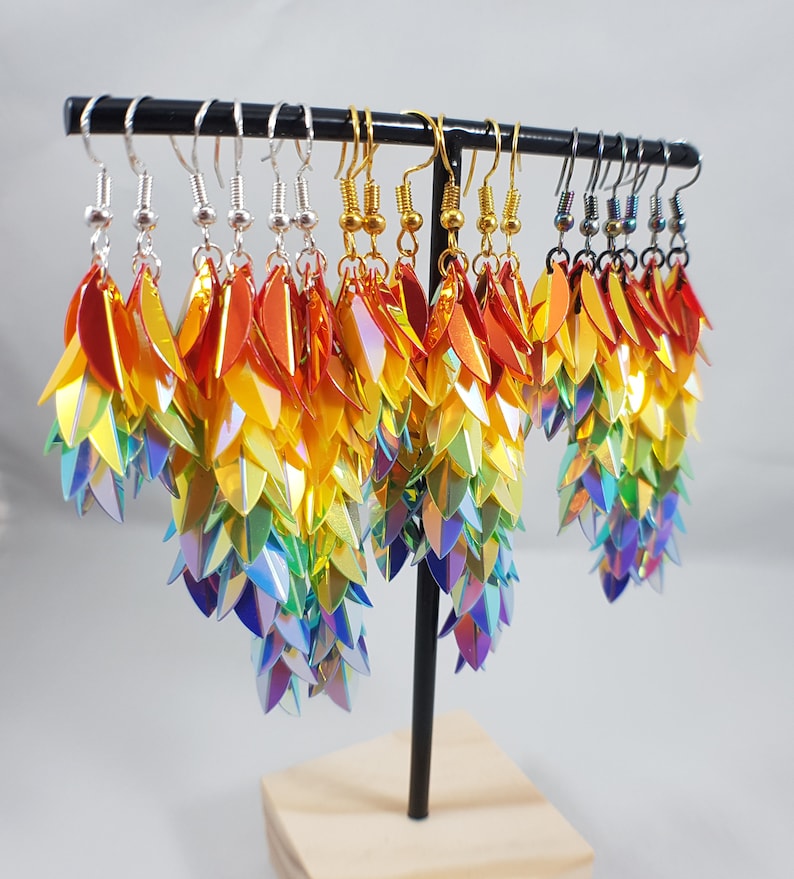 May include: Rainbow iridescent earrings with a scaly design, hanging from a black stand. The earrings are made of small, overlapping scales that create a shimmering effect.