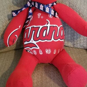 May include: A red teddy bear with a blue ribbon around its neck. The bear's torso features the word "Cardinals" in white script, with "To Baseball" printed below. The bear is a soft toy.