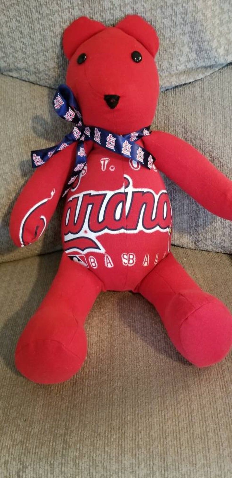 May include: A red teddy bear with a blue ribbon around its neck. The bear's torso features the word "Cardinals" in white script, with "To Baseball" printed below. The bear is a soft toy.