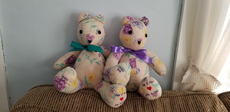 May include: Two handmade teddy bears with floral fabric, each with a ribbon around the neck. One has a teal ribbon, the other a purple ribbon. The bears have embroidered details and personalized text on their feet, including dates and names.