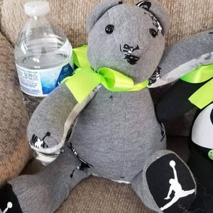 K&ouml;nnte beinhalten: Ein handgefertigter grauer Teddyb&auml;r mit schwarzen Akzenten und einer neongr&uuml;nen Schleife. Der B&auml;r hat schwarz-wei&szlig;e Details an F&uuml;&szlig;en und Ohren. Eine transparente Plastikwasserflasche befindet sich neben dem B&auml;ren.