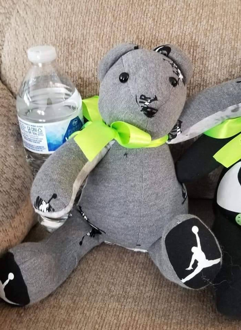 K&ouml;nnte beinhalten: Ein handgefertigter grauer Teddyb&auml;r mit schwarzen Akzenten und einer neongr&uuml;nen Schleife. Der B&auml;r hat schwarz-wei&szlig;e Details an F&uuml;&szlig;en und Ohren. Eine transparente Plastikwasserflasche befindet sich neben dem B&auml;ren.