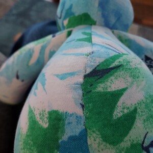 May include: A handmade stuffed animal in the shape of a frog. The frog is made of fabric with a pattern of blue, green, and white abstract shapes. The stitching is visible along the seams.