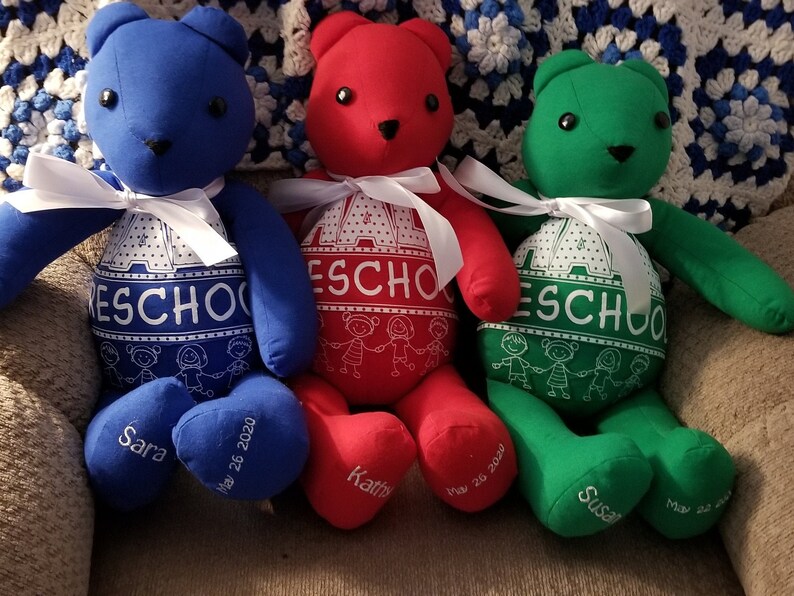 May include: Three colorful handmade teddy bears: blue, red, and green. Each bear has a white ribbon, black button eyes, and the word "Preschool" printed on its belly. The bears' feet are embroidered with names and dates.