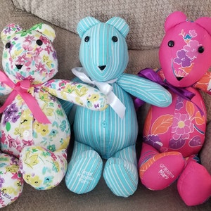 May include: Three handmade teddy bears. One bear is white with a floral pattern and a pink bow. The second bear is light blue with white stripes and a white bow. The third bear is pink with a floral pattern and a purple bow. Each bear has a name embroidered on its foot.