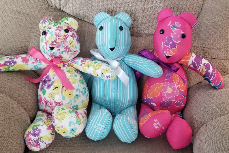 May include: Three handmade teddy bears. One bear is white with a floral pattern and a pink bow. The second bear is light blue with white stripes and a white bow. The third bear is pink with a floral pattern and a purple bow. Each bear has a name embroidered on its foot.