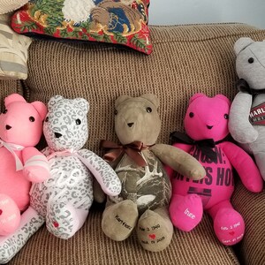 May include: A collection of handmade teddy bears in various colors and patterns, including pink, gray, and camouflage. Some bears have bows, while others feature printed text and dates on their paws. The bears are sitting on a brown couch.