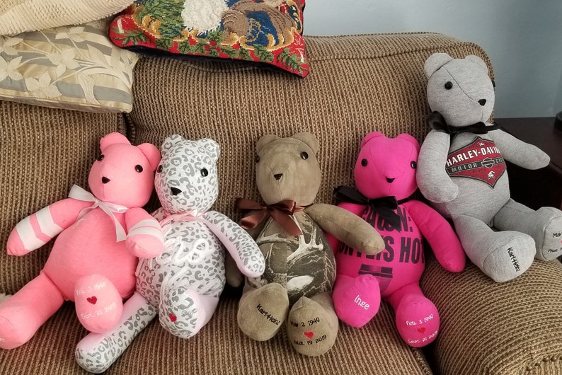 May include: A collection of handmade teddy bears in various colors and patterns, including pink, gray, and camouflage. Some bears have bows, while others feature printed text and dates on their paws. The bears are sitting on a brown couch.