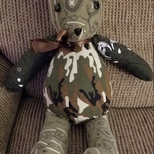 May include: A handmade teddy bear with a camouflage body and paisley patterned head, arms, and legs. The bear has black button eyes and a brown ribbon bow. The bear's foot reads "Mike" and "Dec. 24 1951".