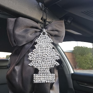 May include: A car air freshener in the shape of a tree, encrusted with sparkling rhinestones, hangs from a black satin bow. The bow is attached to the interior of a car. The air freshener adds a touch of glamour to the vehicle's interior.