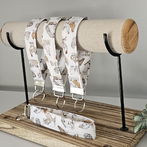 May include: A display of several white lanyards with a chicken and coop pattern, hanging from a cylindrical, fabric-covered stand. A matching wristlet lanyard rests below. The stand is supported by black metal legs and a wooden base.