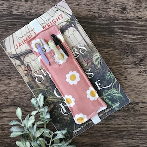 May include: A pink fabric pen holder with a daisy pattern, holding several pens, rests on top of a book. The book has a floral design and the author's name, Jaime Wright, is visible. A sprig of greenery is in the foreground.