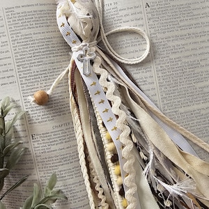 May include: A handmade tassel bookmark with a cross charm. The bookmark features a wooden bead, cream-colored ribbons, and a silver cross, resting on an open book with text.