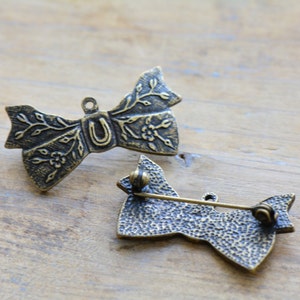 Puede incluir: Dos broches en forma de lazo de color bronce. Los broches presentan diseños florales y de hojas, con una herradura en el centro de uno. Los broches tienen un alfiler en la parte posterior para sujetarlos.