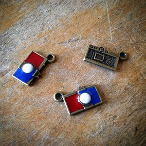 May include: Three metal charms with enamel paint. One charm is a blue and red rectangle with a white circle in the center. Another charm is a blue and red rectangle with a white circle in the center. The third charm is a brown rectangle with a textured surface and a white circle in the center.