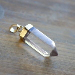 Może przedstawiać: Wisiorek z przezroczystego kryształu kwarcowego z pozłacanym metalowym zapięciem. Wisiorek ma kształt długiej, spiczastej kropli.